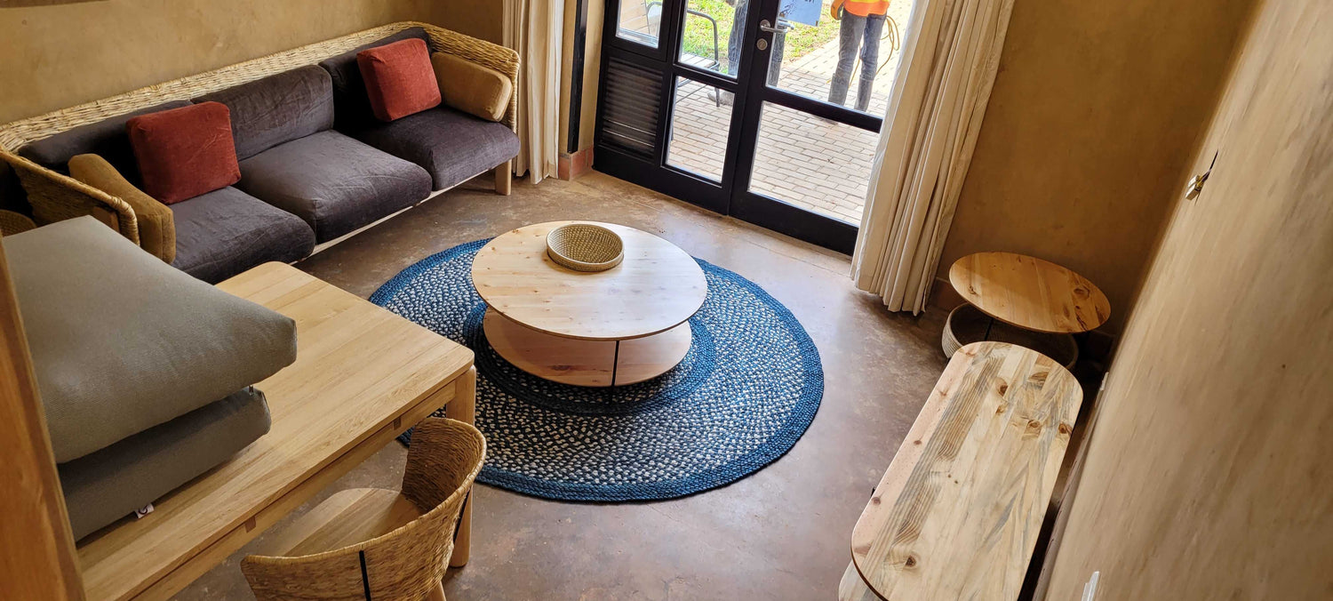 Cozy living room featuring a natural woven sofa with plush cushions, a wooden coffee table with storage compartments, a round woven rug, and large windows with curtains allowing natural light.
