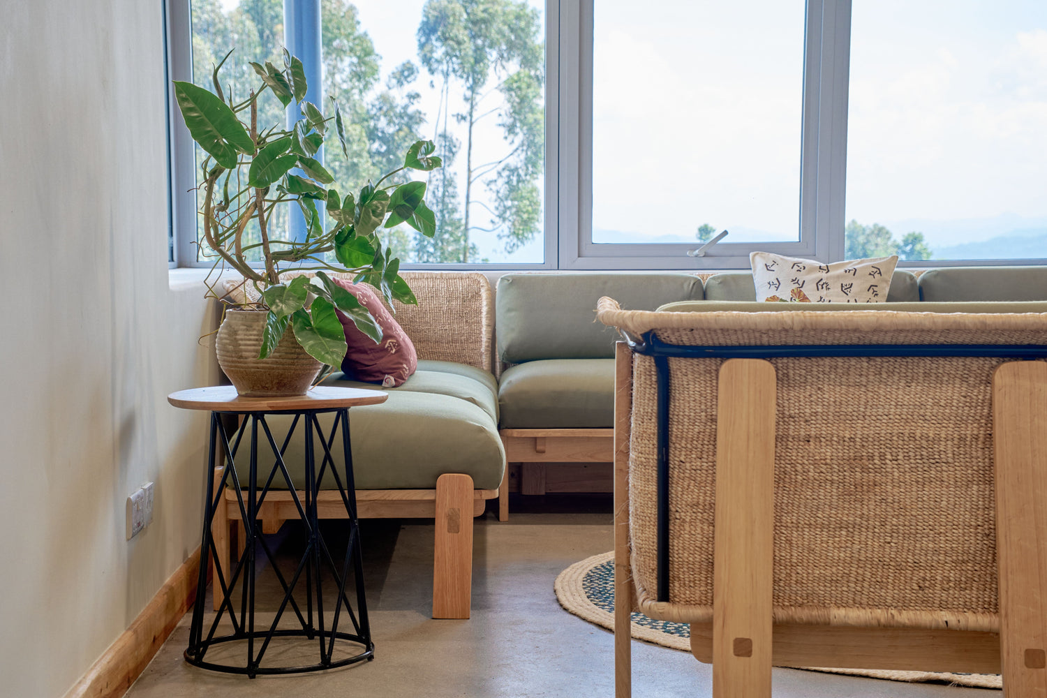 Bright seating area with a green cushioned sectional sofa framed by woven natural-fiber accents, a round wooden side table with a black metal base, and a potted plant.