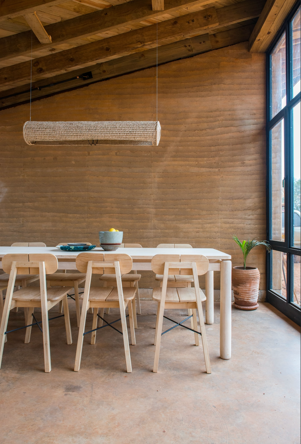 Modern dining setup with a sleek wooden table, matching chairs, and a woven pendant light, surrounded by warm-toned walls and large glass windows with natural light.